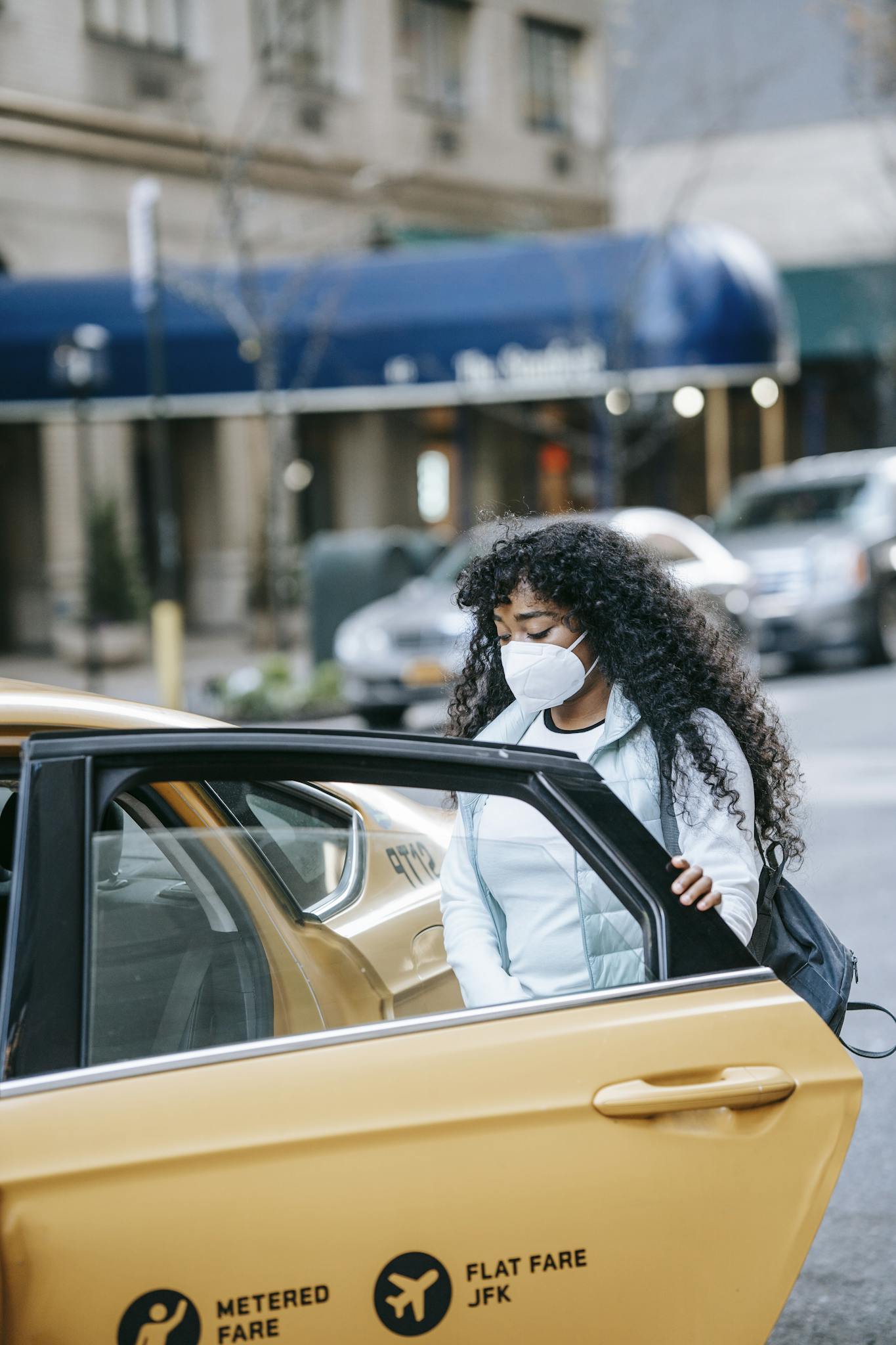Calm young African American millennial with long curly hair in casual outfit and protective mask getting into yellow cab on city street